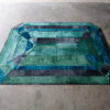 Luxury designer rug featuring green, teal, blue, and black rectangles in a stepped octagonal pattern on concrete flooring.