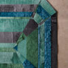 A close-up of a luxury designer rug, Custom Emerald 02709, with geometric patterns in greens, blues, and grays on a beige concrete floor.