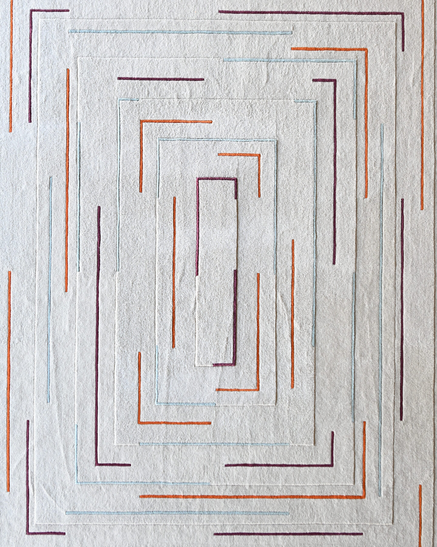 A luxury designer rug with concentric rectangles in purple, orange, and blue on a textured light gray background.