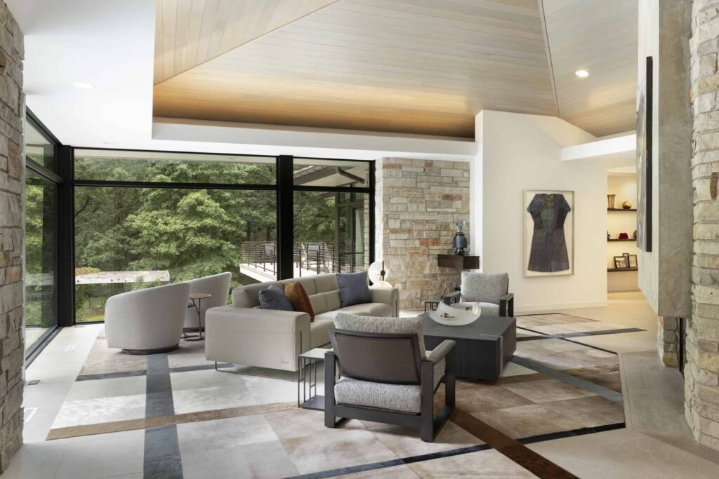 Modern living room with luxury designer rug, neutral furniture, stone walls, and forest views.