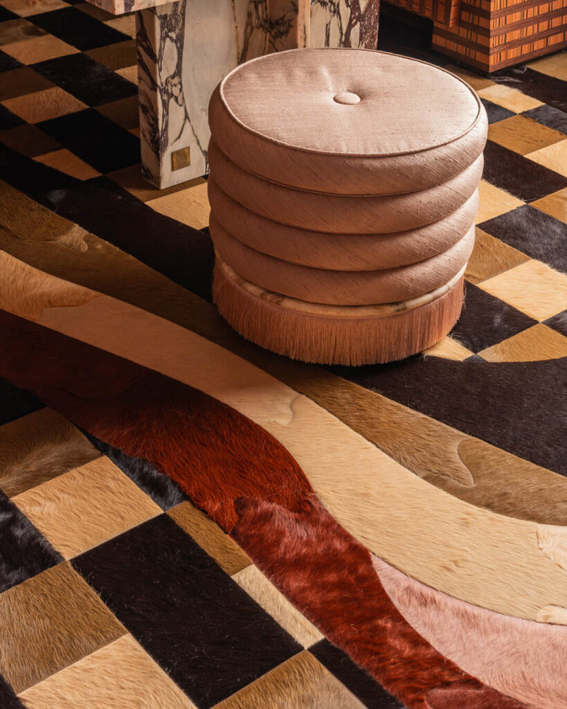 A round beige ottoman sits on a luxury designer rug with geometric patterns and earth tones.