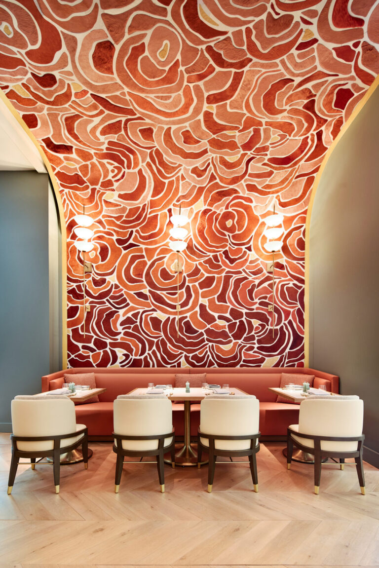 A restaurant booth with luxury custom rugs, beige chairs, and a red bench, set by abstract rose-patterned walls.