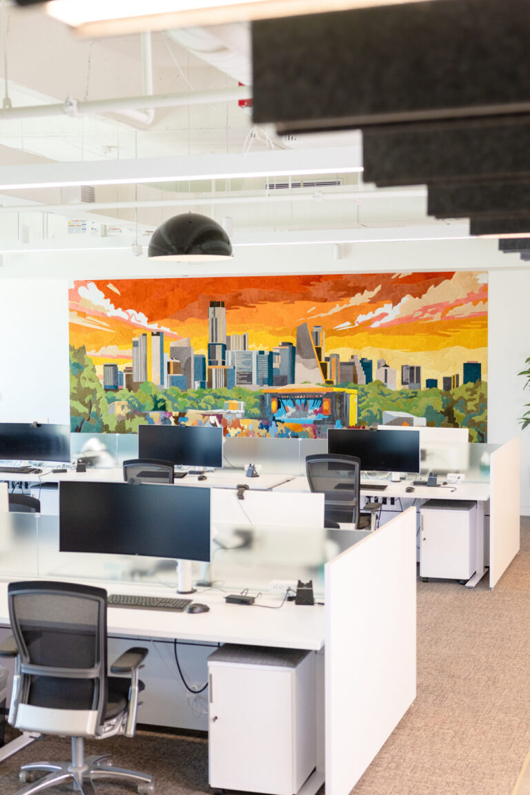 Open-plan office with designer rugs and white desks, computer monitors, black chairs, and cityscape mural.