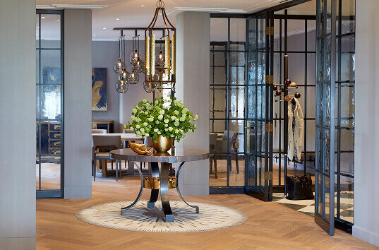 A round table on a luxury designer rug in a glass entryway with pendant lights above.