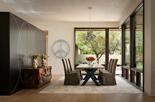 A modern dining room with a rectangular table atop a luxury designer rug, floor-to-ceiling windows, and peace symbol wall art.