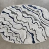 The Submerge Hide 27443 luxury designer rug features an oval with wavy lines in white, gray, and dark blue on a premium neutral background.