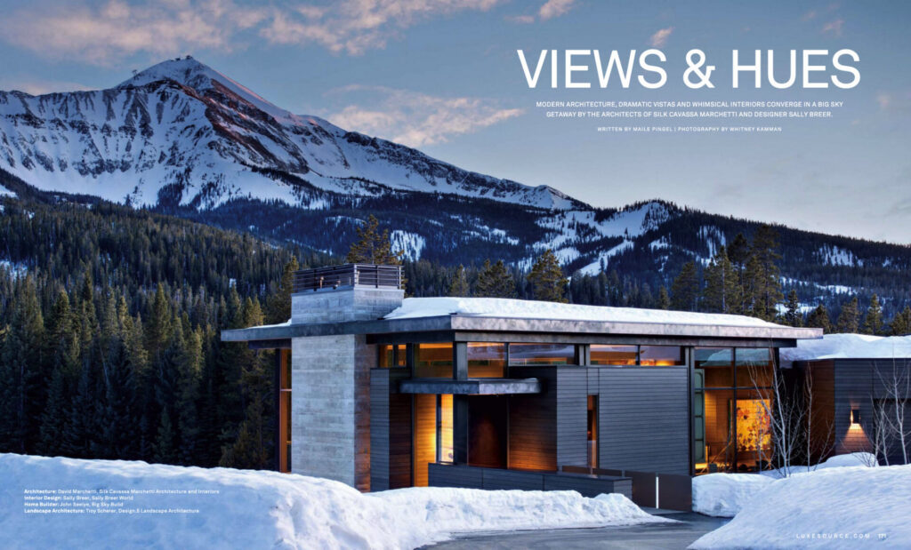 A modern house with large windows and luxury designer rugs overlooks a snowy landscape at dusk.