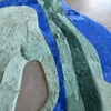Luxury designer rug features abstract blue, green, and beige layers on smooth gray hide.
