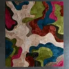 Luxury designer square rug with abstract shapes in beige, green, blue, pink, and red on concrete floor.