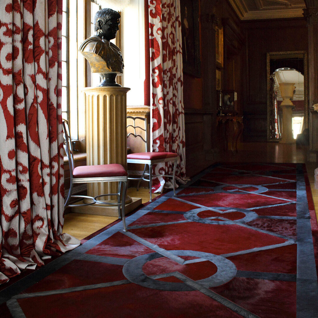 A hallway with luxury custom red and black designer rug, classic bust, and patterned curtains.