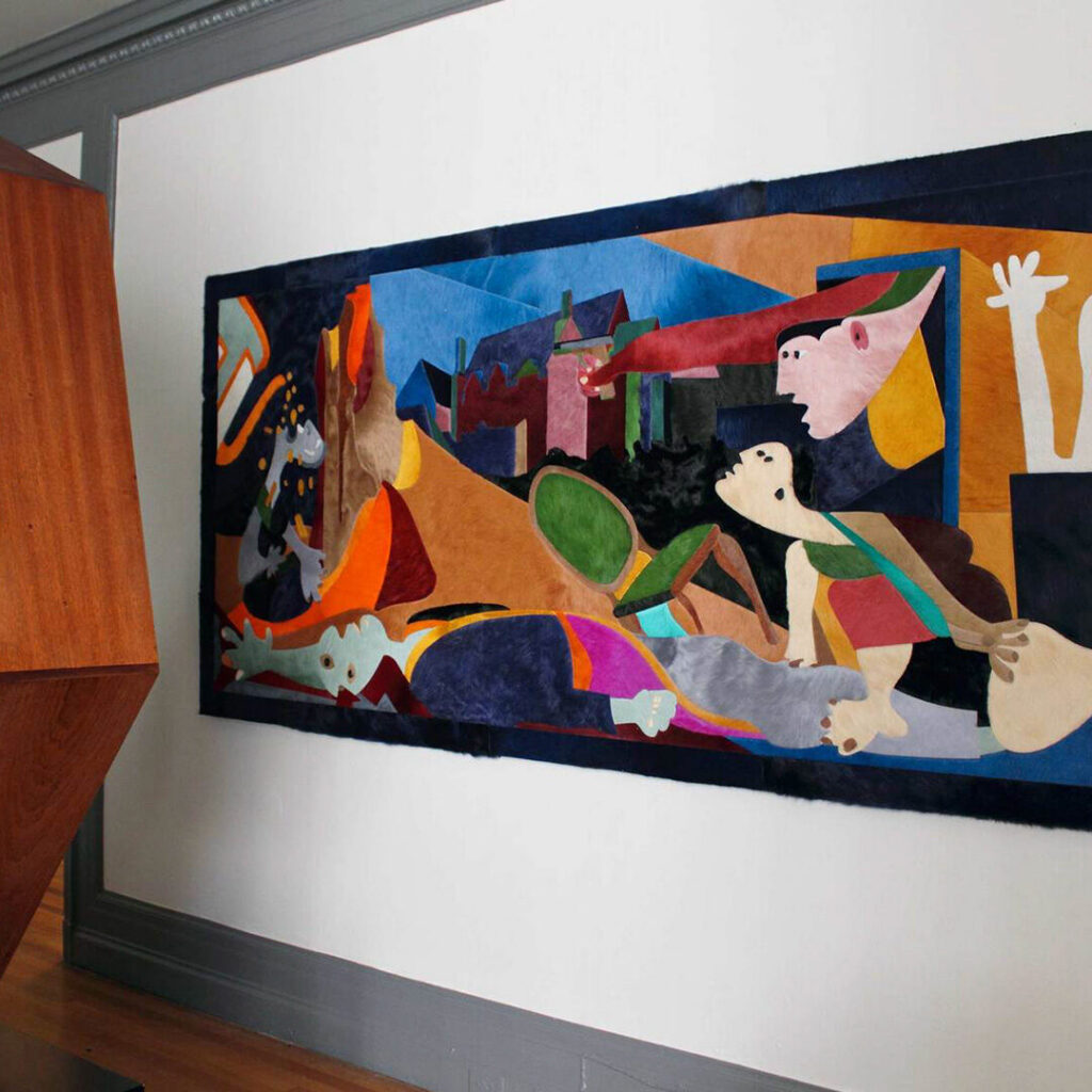 A luxury designer rug tapestry with geometric and figurative shapes adorns a white wall beside angular wood.